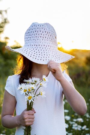 a beautiful little girl with daisies covers her face with a hat.の写真素材