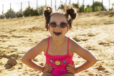 a beautiful girl in a pink swimsuit and glasses is sitting on a sandy beach and laughing.の写真素材