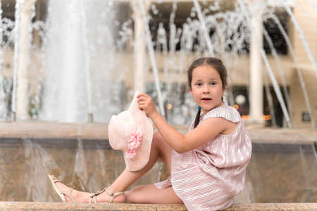 a beautiful little girl is sitting on a fountain and holding a hat.の写真素材