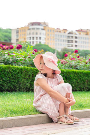 a beautiful little girl in a hat sits on the curb and looks away.の写真素材