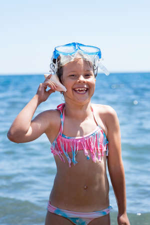 beautiful little girl in a swimsuit listening to a seashell on the beach.の写真素材