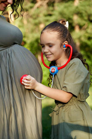 Pregnant woman with her daughter. Little girl excited about her coming sibling - listening to mothers tummyの写真素材