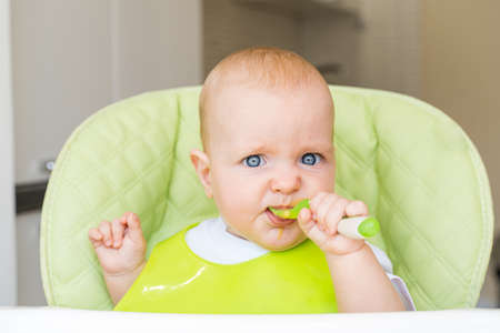 a small child with blue eyes and blond hair sits in a high chair and eats the first bait. Healthy diet. Baby 6 months.の写真素材