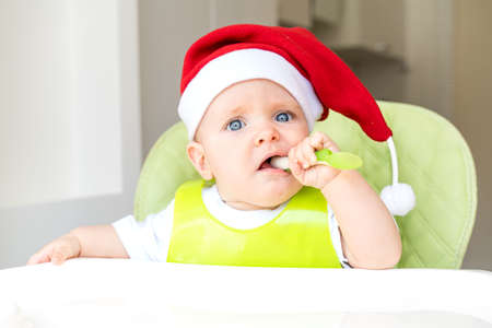 a baby in a Santa Claus hat is sitting in a high chair and eating with a spoonの写真素材