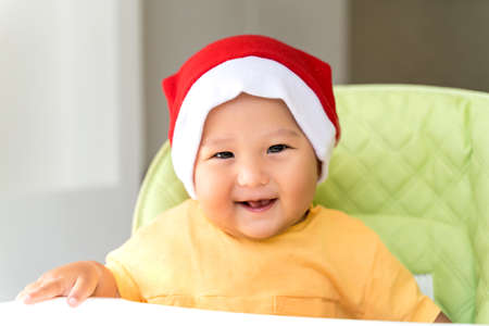 asian baby in a Santa Claus hat is sitting in a high chair and eating with a spoonの写真素材