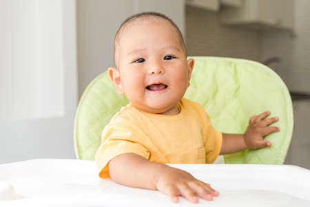 asian baby is sitting in a high chair and eating with a spoonの写真素材