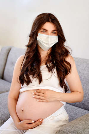 Young beautiful pregnant woman in medical surgical mask and white t-shirt quarantined at home by the window. Coronavirus, virusの写真素材