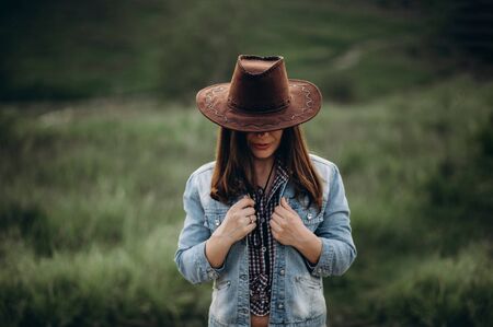 Girl alone in the field in the evening holds on to a denim jacket as if frozen on the head cowboy hat outsideの写真素材