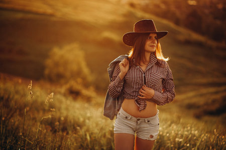 Happy young cowgirl standing at farm fence. Woman wearing blouse, cowboy hat. Countryside, farming,...