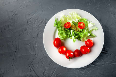 greens of lettuce and cherry tomatoes in the form and form of an emoji on a white plate on a gray backgroundの写真素材