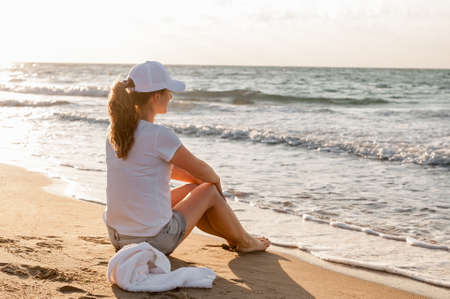 the athlete sits on the beach after a jog and looks at the sunset. Girl in white T-shirt and capの写真素材