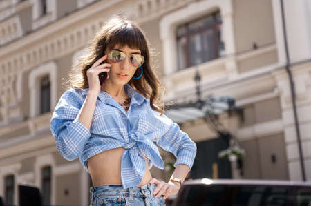 Outdoor portrait of yong beautiful happy smiling woman wearing stylish sunglasses, blue jeans blouse, blue mom jeans, with small quilted bag. Model posin in street of european city. Copy spaceの写真素材