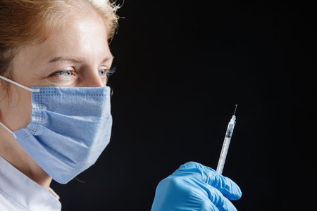 Portrait of a doctor or nurse in a medical uniform and protective blue mask with a syringe in his...