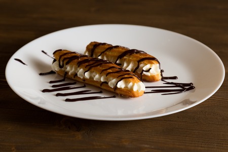 Eclairs with a delicate cream inside, covered with black chocolate on a white plate on a brown wooden table. Homemade recipe bookの写真素材
