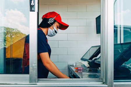 Bangkok, Thailand - August 4, 2020 : fast food cashier in drive thru service waring hygiene face mask to protect coronavirus pandemic or covid-19 virus outbreak working on counter at the station.のeditorial素材
