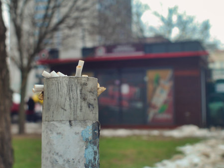 Close-up of a weathered metal public ashtray filled with discarded cigarette filters. This editorial urban shot captures themes of nicotine addiction, city pollution, and unhealthy social habits.の写真素材