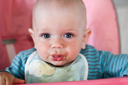 Beautiful baby eats porridge from mom's hand. He is sitting on a pink children's chair