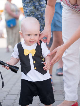 Beautiful toddler walking with his mother on a city holiday. Kid stylishly dressed butterfly and cap. Boy holding a flashlight.の写真素材