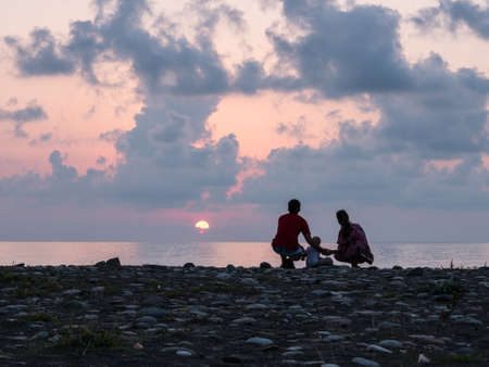 Happy family - father, mother, baby son see sunset sea surf on black sand beach. Active parents and people outdoor activity on tropical summer vacations with children.の写真素材