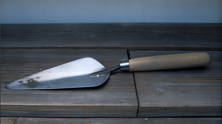 A metal trowel with a wooden handle rests on a dark wooden surface. The trowel shows signs of use.の写真素材