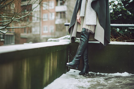Close-up of a woman's legs in black boots on a snowy urban scene. She is wearing a beige dress and a grey coat.の写真素材