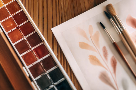 Close-up overhead view of watercolor paints, brushes, and a painting in progress on a wooden...