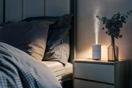 A white humidifier sits on a bedside table next to a bed with striped bedding, softly illuminating the room.の写真素材