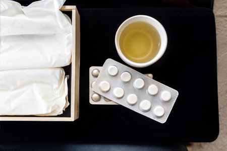 Top view of blister packs of pills, a cup of tea, and tissues in a wooden box on a black surface.の写真素材