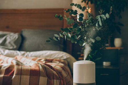 White humidifier with eucalyptus in a bedroom with soft lighting and a plaid blanket on the bed.の写真素材
