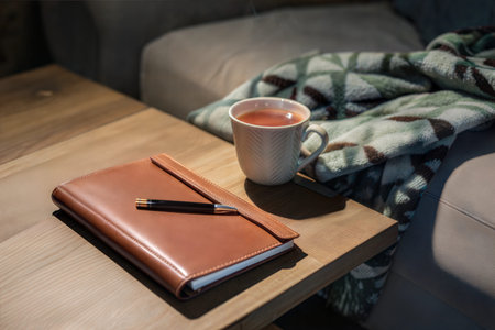 A warm cup of tea sits on a wooden table next to a leather journal and pen. A cozy blanket is...