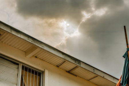 Low angle view of a house roofline against a cloudy sky, with a glimpse of sun. A fabric hangs on a...