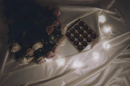 A bouquet of roses sits next to a box of chocolates on a silk fabric with fairy lights.の写真素材