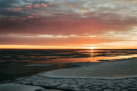 Vibrant sunset colors paint the sky over a partially frozen lake, with ice floes and patches of open...