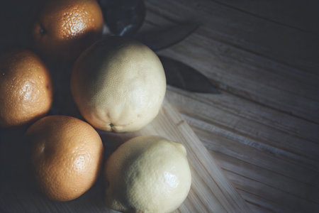 Close-up of grapefruits and a lemon on a wooden cutting board, with a knife in the background. Dark and moody lighting.の写真素材
