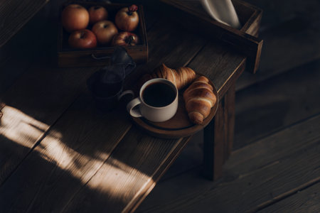 A rustic scene of coffee, croissants, and apples on a wooden table, bathed in sunlight.の写真素材