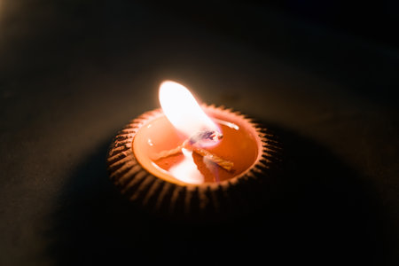 A close-up shot of a candle burning brightly in the dark, casting a warm glow. The candle is in a small, decorative holder.の写真素材