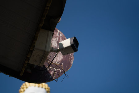 A white security camera mounted under a roof eave against a clear blue sky, with a glimpse of a...