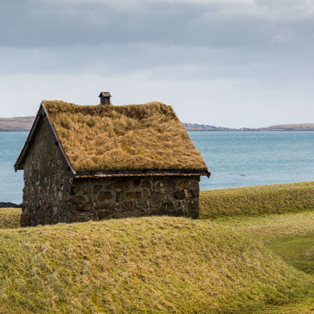 Traditional hut with a green grass roof with a backdrop of the Atlantic ocean and an islandの写真素材