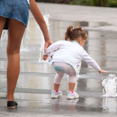 A young mother with beautiful legs tries to take away the funny little girl from the fountain, but her daughter wants to playの写真素材