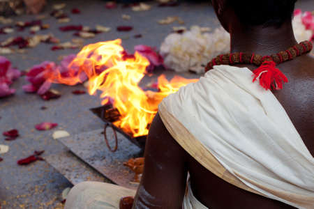 Hindu is sitting near a fire wearing traditional holiday clothes; on the ground flower petals have been stripped and a garland of flowers liesの写真素材