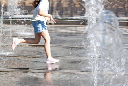 teenager girl joyfully runs through the water among the spray of fountains in a summer sunny city parkの写真素材