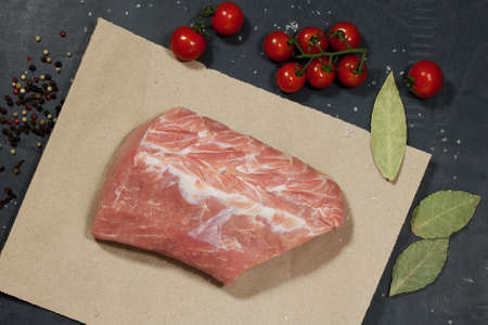 piece of meat lies on paper on a black table. there is also a branch of cherry tomatoes and spices for making delicious homemade food.の写真素材
