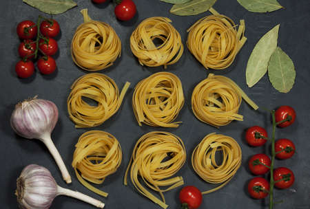 on a black table are raw twisted noodles, cherry tomato twigs, garlic, bay leaf and pepper. everything is prepared for cooking a delicious lunch for several portionsの写真素材