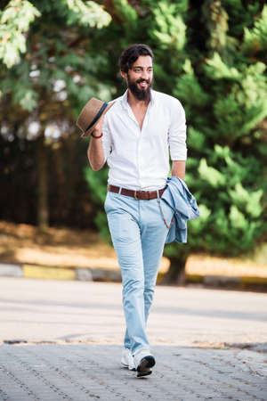 Stylish man walking on a street, with mustache and beard, holding hat and a vest, wearing blue pants and a white shirt, in front of a natural green backgroundの写真素材