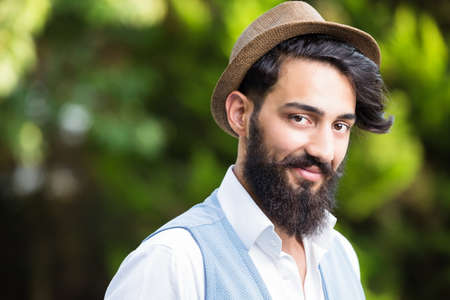 Headshot stylish man with beard and mustache wearing brown line, white shirt and blue vest in front of a natural green tree out of focus backgroundの写真素材