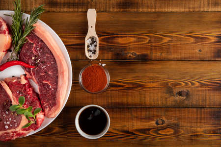 Chunks of raw meat on a white plate. Chili pepper, rosemary, spices. Wooden table. Top view.の写真素材