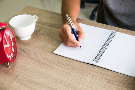 Closeup of male right hand writing text on blank notebook on wooden desk in office.