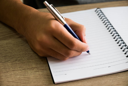 Closeup of male right hand writing text on blank notebook on wooden desk in office.