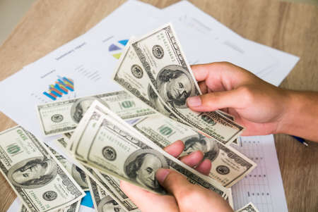 Closeup of Asian male hands counting bunch of dollars banknotes with business charts background for business, finance, tax and people conceptの写真素材