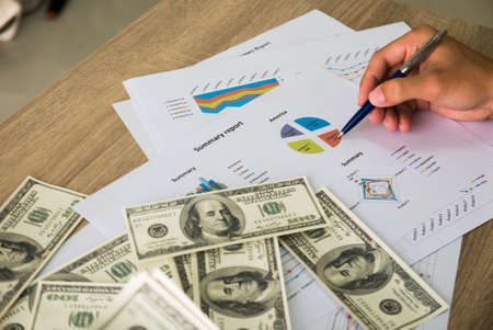 Closeup of Asian male left hand counting bunch of dollars banknotes and his right hand hold ball pen points on business charts background for business, finance, tax and people conceptの写真素材